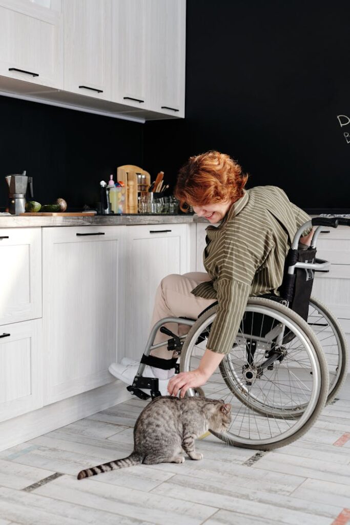 A woman in a wheelchair smiles while petting a tabby cat in a modern kitchen.